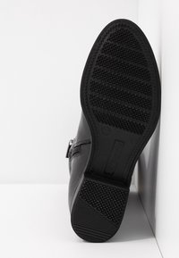 Svart lädersula på skyddssko med texturerad sula, storlek 37, lutad mot vit hörnvägg och golv som bakgrund.