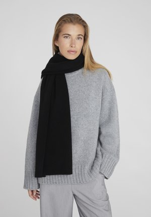 Pull en maille gris avec un ourlet côtelé, assorti à une longue écharpe noire. L'écharpe présente une texture lisse, et les deux vêtements ont une coupe décontractée.