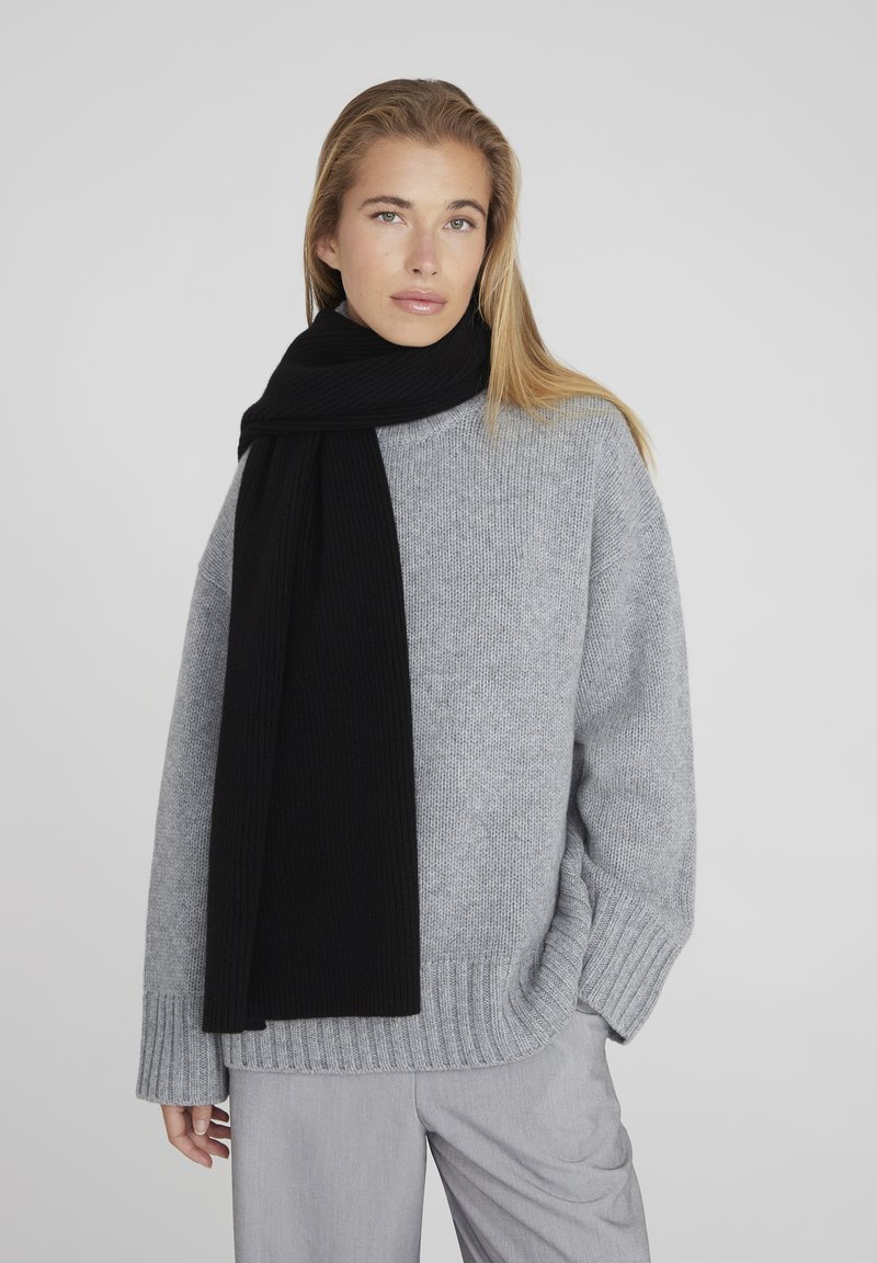 Pull en maille gris avec un ourlet côtelé, assorti à une longue écharpe noire. L'écharpe présente une texture lisse, et les deux vêtements ont une coupe décontractée.