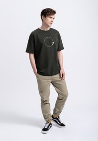 Donkergroene t-shirt met korte mouwen en een cirkelvormige grafiek, gecombineerd met beige broek en zwarte sneakers. Glad stof en relaxte pasvorm.
