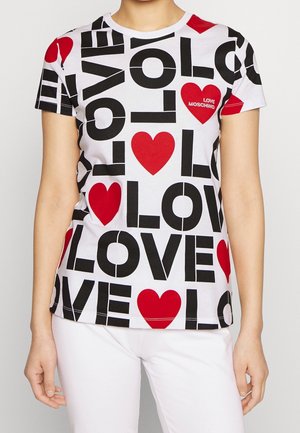 Henkilö, jolla on valkoinen T-paita, jossa on lihavoitu musta "LOVE" -teksti ja punaisia sydämenmuotoja, "LOVE MOSCHINO" painettuna sydänkuvioon. Valkoiset housut.
