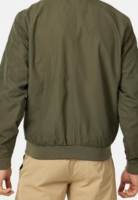 Homme portant une veste bomber vert olive et un pantalon beige, vu de dos avec les mains détendues le long du corps.