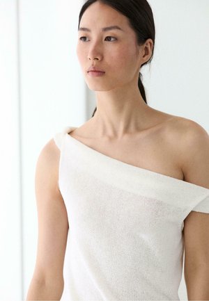 Giovane donna con capelli scuri in un top bianco monospalla in maglia che guarda di lato contro uno sfondo luminoso e minimalista.