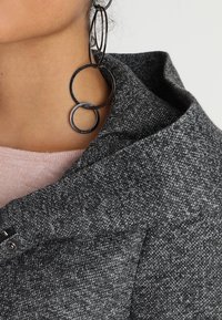 Harmaasta teksturoidusta kankaasta valmistettu vaatekappale, jossa on taiteltu kaulus, napitus ja suuret pyöreät mustat korvakorut lisäyksinä.