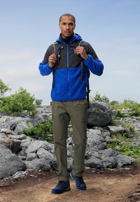 Blå och grå zip-up jacka med huva, i kombination med olivfärgade cargobyxor och blå vandringsskor, mot en stenig utomhusbakgrund.