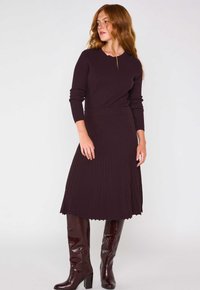 Abito lungo, a coste, di un viola scuro con corpetto aderente e gonna svasata. Abbinato a stivali al ginocchio, lucidi, di color bordeaux. Tessuto texturizzato.