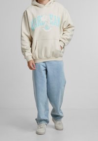Ljust beige hoodie med "KARL KAN" i turkoss grafisk tryck, tillsammans med ljusblå baggy jeans och vita sneakers.