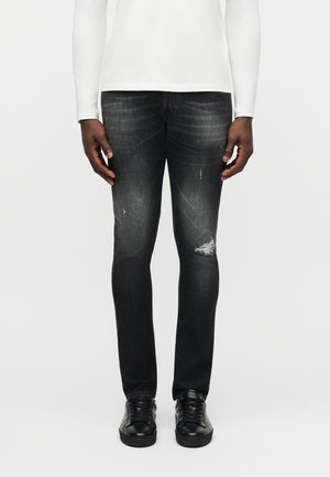 Muž v čiernych opotrebovaných úzkych džínsoch s čiernymi teniskami a bielou košeľou s dlhým rukávom stojaci pred jednoduchým pozadím.