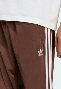 Barna sportnadrág sima anyagból, fehér oldalcsíkokkal és hímzett Adidas logóval a bal lábon.