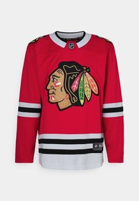 Maglia da hockey rossa con un grande logo indiano americano sul davanti, strisce nere e bianche sulle maniche e un collo rotondo.