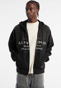 Schwarze Zip-Hoodie mit strukturiertem Finish, ausgestattet mit einer Doppellage-Kapuze und bedrucktem Logo auf der Vorderseite. Darunter befindet sich ein weißes T-Shirt.