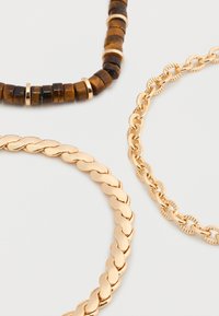 Trois bracelets : un avec des perles de pierre brunes et des anneaux dorés, un gros maillon lisse en or, et une fine chaîne en or texturée sur fond blanc.