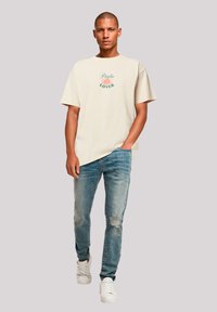 Beige katoenen t-shirt met groene "Pasta Lover" graphic, gecombineerd met versleten blauwe spijkerbroek en witte sneakers. Eenvoudig en casual ontwerp.