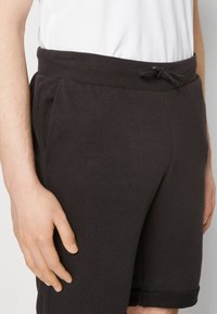 Shorts en coton noir avec une taille élastique, fermeture à cordon et poches latérales. Le tissu a une texture lisse, et l'ourlet est retroussé.