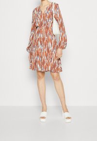 Vestido de color marrón y azul con estampado, de mangas largas y abullonadas, escote en V, talle fruncido y falda acampanada, combinado con sandalias de plataforma blancas.