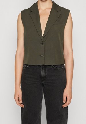 Ærmeløs olivengrøn cropped blazer med to knapper kombineret med sorte højtaljede jeans på en person, der står mod en ensfarvet baggrund.