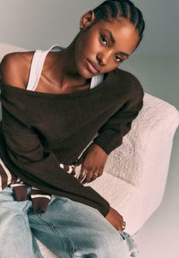 Bruine oversized trui met geribbelde manchetten, van de schouder gedragen, gecombineerd met blauwe spijkerbroek. Zachte textuur en gelaagd met een witte tanktop.