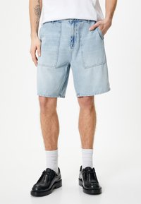 Lys blå denimshorts med afslappet pasform, der har to store forlommer og en frynset kant. Sammen med sorte sko og hvide sokker.