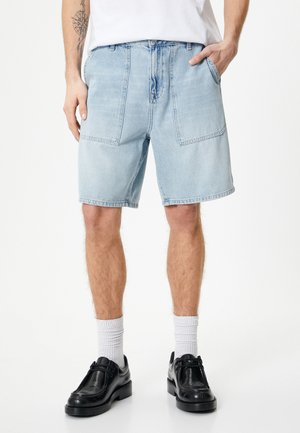 Lys blå denimshorts med afslappet pasform, der har to store forlommer og en frynset kant. Sammen med sorte sko og hvide sokker.