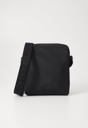 Bolso bandolera negro de nailon con una textura suave, que presenta un logo "CK" y una correa ajustable con el nombre de la marca tejido.