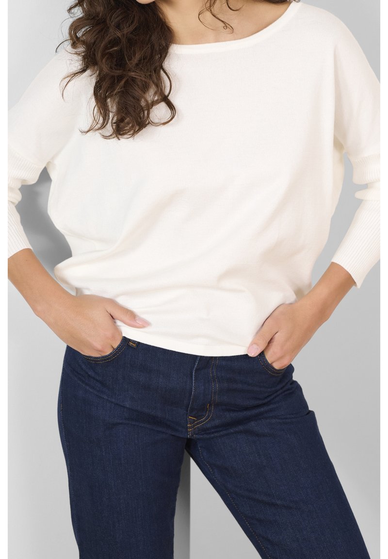 Femme aux cheveux bruns bouclés portant un pull blanc ample et un jean bleu foncé, les mains dans les poches, devant un fond uni.