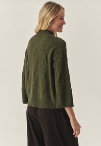 Adīts zaļš džemperis ar augstu apkakli, strukturētiem rakstiem un plašām piedurknēm. Nēsāts kopā ar tumšām biksēm, izceļot brīvu piegriezumu un ribotu apakšmali.