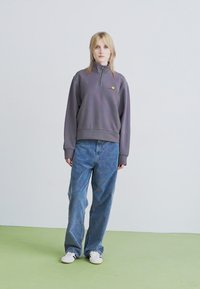 Mujer joven de pie, vestida con un sudadero gris de media cremallera, jeans azules holgados y zapatillas blancas, contra un fondo pálido y sencillo.