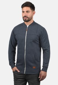 Tmavomodrá mikina na zips s okrúhlym golierom, prednými vreckami a koženým akcentom na leme. Vyrobená z mäkkej, textúrovanej látky.