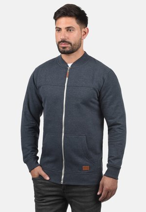 Sweatshirt zippé bleu foncé avec un col rond, des poches avant et un accent en cuir sur l'ourlet. Fabriqué en tissu doux et texturé.