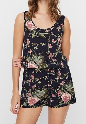 Kvinde iført navyblå ærmeløs romper med pink og grønt blomsterprint, står med den ene arm krydset over torsoen.
