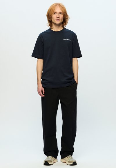 Marineblå bomuld T-shirt med korte ærmer, med et lille hvidt logo på venstre bryst; parret med sorte bukser med striber og sneakers.