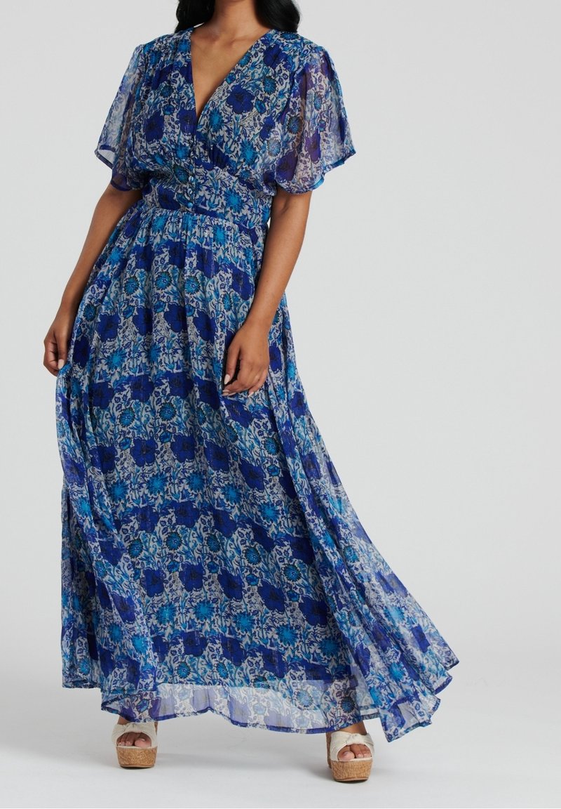 Robe maxi en tissu bleu transparent avec des motifs floraux. Présente un décolleté en V profond, des manches courtes évasées et une taille cintrée. Portée avec des sandales compensées.