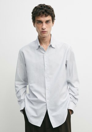 Junger Mann mit lockigem Haar, der ein hellgrau-weiß gestreiftes Hemd mit Knöpfen trägt und die Hände in den Taschen hat, vor einem schlichten Hintergrund.