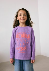 Lavendli pikad varrukad särk, millel on koralli graafika bubble tee tassist; sisaldab teksti "Magus nagu BUBBLE TEA" ja "ERILINE VERSIOON."