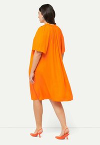 Vestido corto naranja de manga corta con escote fruncido, silueta fluida y textura suave. Combinado con zapatos de tacón alto naranjas.