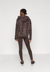 Mikina s kapucňou so vzorom leopardej potlače v tmavo hnedej a béžovej farbe, kombinovaná s hnedými legínami. Má klokanie vrecko a voľnejší strih s kapucňou.