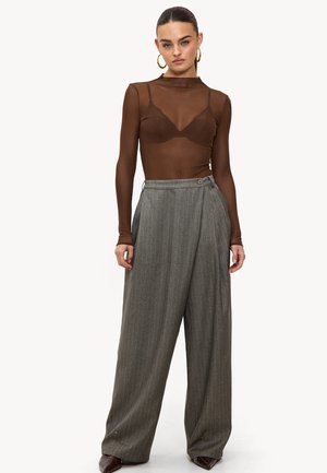 Femme portant un haut en transparent marron à manches longues avec un soutien-gorge visible, un pantalon large en gris à rayures, des boucles d'oreilles en or, et des chaussures pointues marron.