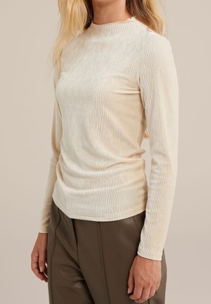 Nainen, joka pukeutunut kermanvalkoiseen, teksturoituun pitkähihaiseen toppiin, joka on sullottu korkeavyötäröisten oliivinvihreiden housujen sisään, seisomassa neutraalia taustaa vasten.