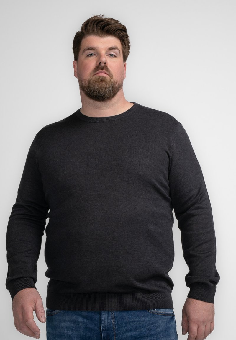 Pulover din tricot de culoare gri închis, cu un decolteu rotund și mâneci lungi, având manșete și tiv cu o textură ribbed. Asortat cu blugi albaștri.