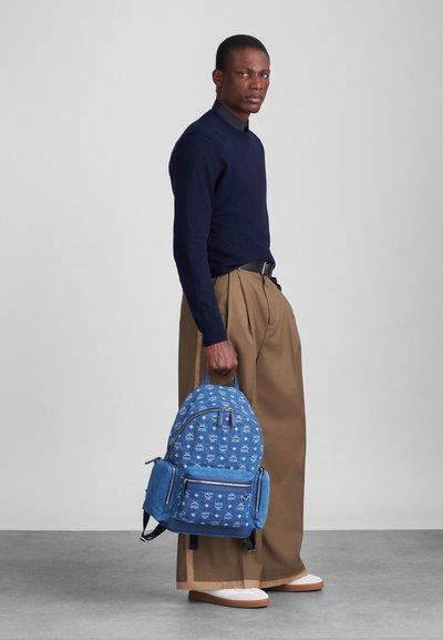 Μπλε τσάντα πλάτης από τζιν με λευκά γραφικά σχέδια, διαθέτοντας φερμουάρ και τσέπες; φορεμένη από ένα άτομο με σκούρα παντελόνια και ναυτικό πουλόβερ.