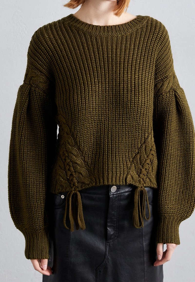 Pull en maille épaisse vert olive avec manches bouffantes, détails en maille torsadée et liens à l’ourlet, porté avec une jupe en cuir noire.