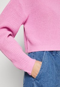 Rosa, kurz geschnittenen Strickpullover mit strukturiertem Muster und langen Ärmeln, kombiniert mit blauen, hoch taillierten Jeans.