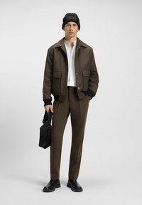 Brown textured jacket with black cuffs and two front pockets, white shirt, brown pants, and black bag. Complements with a black beanie.