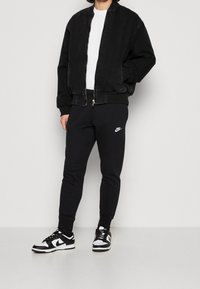 Persona vestida con chaqueta bomber negra, camisa blanca, pantalones de chándal negros de Nike y zapatillas negras y blancas de Nike, de pie contra una pared lisa.