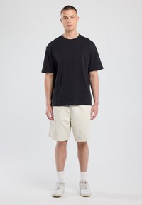 Camiseta de algodón negra de manga corta, combinada con pantalones cortos beige claro. Zapatillas blancas con cordones texturizados y calcetines blancos acanalados.
