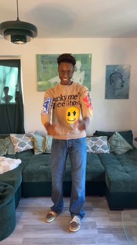 Tânăr zâmbitor în interior, purtând un hanorac bej cu față zâmbitoare galbenă și blugi albaștri, stând în fața unui sofa verde și a unei lucrări de artă pe perete.