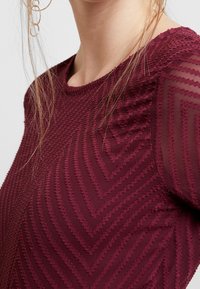 Burgunderfarbenes Oberteil mit einem strukturierten Zickzackmuster, das über Netzärmel verfügt. Runder Ausschnitt, figurbetonte Passform und aufwendig gestickte Details.