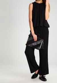 Femme en top sans manches noir et pantalon large portant une pochette noire à rayures diagonales, avec des mocassins à franges noirs.