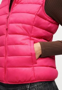 Χέρι στην τσέπη ενός φωτεινού ροζ καπιτονέ γιλέκου που φοριέται πάνω από καφέ ριγέ μακρυμάνικο μπλουζάκι και σκοτεινά τζιν με ξεβαμμένο ύφασμα.