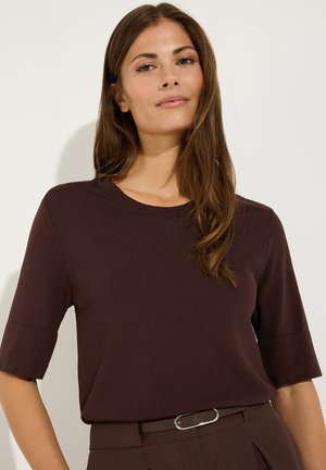 Femme aux longs cheveux bruns portant un haut à manches courtes marron foncé et un pantalon assorti avec une ceinture fine, debout devant un fond clair uni.